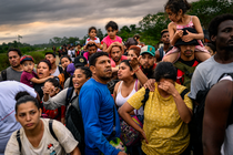 photo of crowded line of people wearing backpacks, several carrying a child on their shoulders, one woman with hand to forehead