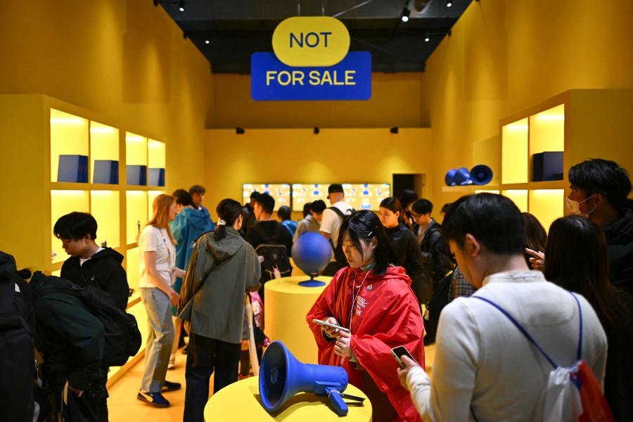 People walk through an exhibit designed like a store, looking at blue-colored items on yellow shelves, beneath a sign reading "Not for sale."