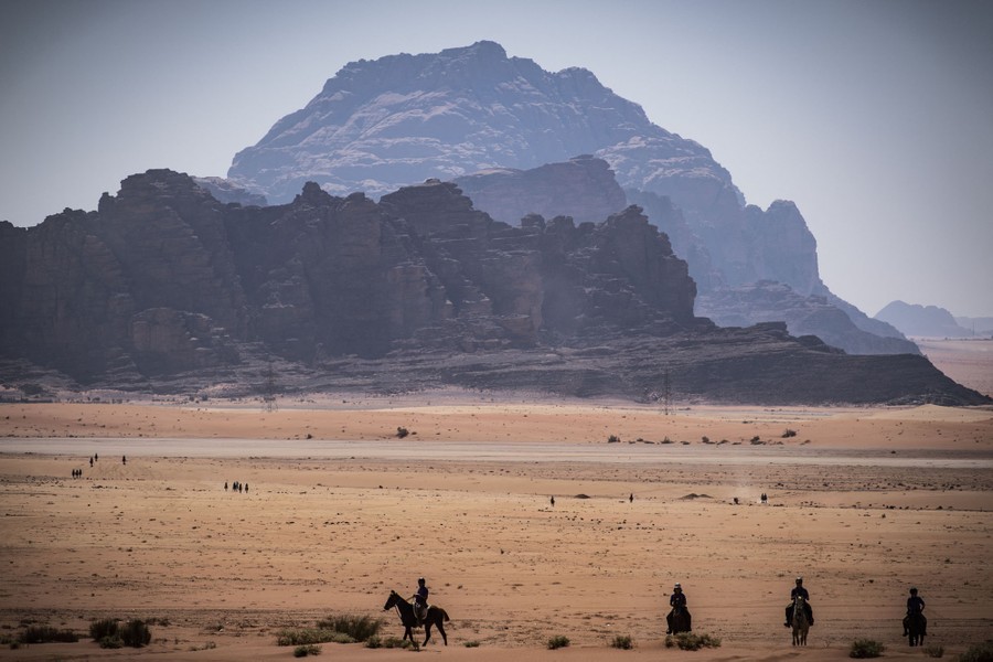 Several people ride horses in a desert with rocky mountains in the background.