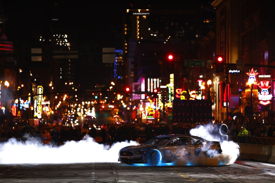 A race car is driven in the middle of a city street at night, sending smoke up from its tires.