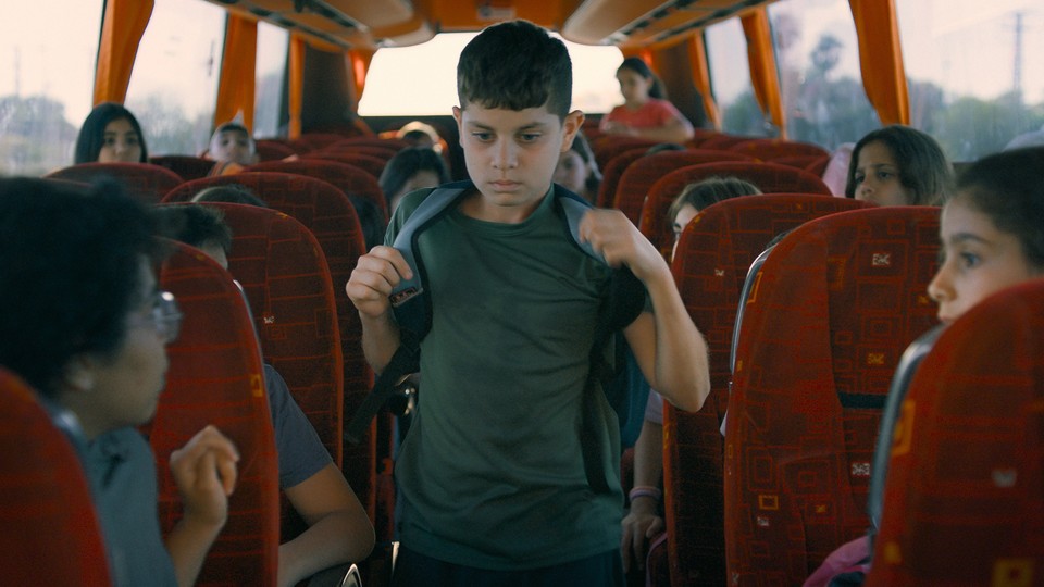 Color photograph of a young boy wearing a backpack walking down the aisle of a bus as sitting children stare at him.