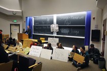 A professor stands at a lecture podium, while protesters sit alongside him holding signs. 