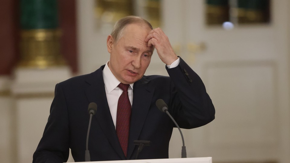 Vladimir Putin stands at a lectern, looking downward and holding his hand up to his head.