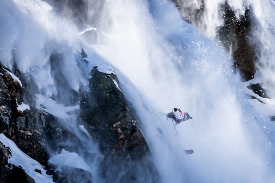 A snowboarder descends a steep, rocky, snow-covered mountain during a competition.