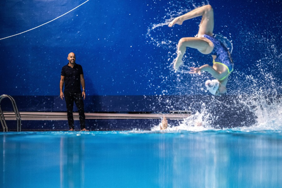 A person stands alongside a pool as a swimmer leaps out of the water, splashing and tumbling.