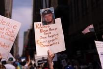 A hand holds up a sign that reads “Release Kilmar Abrego Garcia / Cease targeting Immigrant Communities” at a protest where others wave signs and upside-down American flags