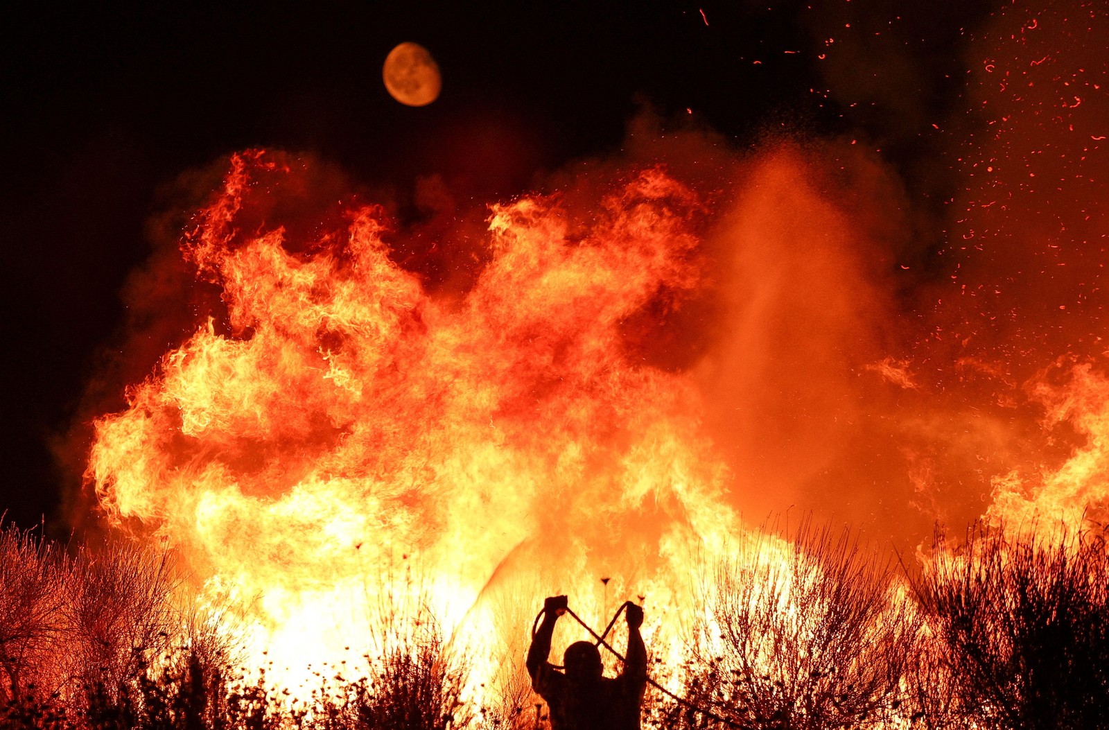 The moon is visible in the sky above a large wildfire, with a silhouetted firefighter below.
