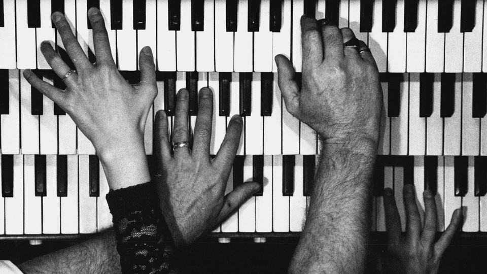 Black-and-white photo of many different hands playing three stacked sets of piano keys.