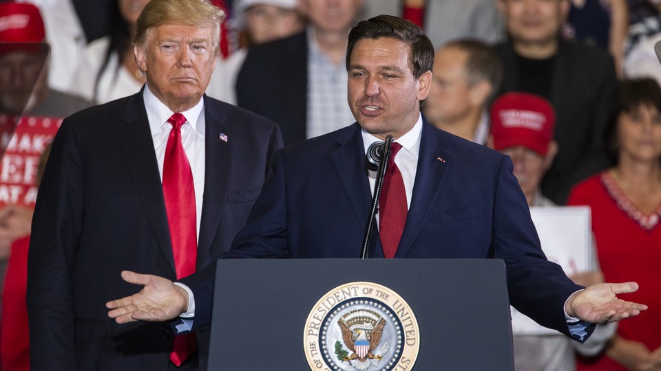Ron DeSantis stands at a podium, his arms spread open. Donald Trump stands just behind him, on the left.