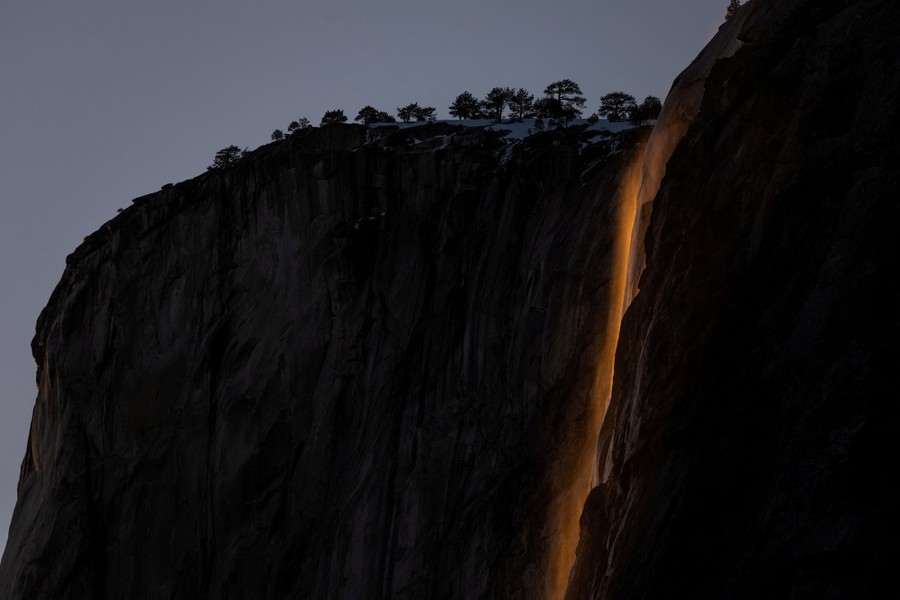 A tall waterfall on a steep, rocky mountain is lit by low sunlight, causing it to glow against the darker background.