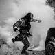 A black-and-white photo of an ICE agent between two cars and a cloud of tear gas in the background
