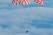 A gumdrop-shaped spacecraft, charred from the heat of reentry, coasts through blue sky beneath a trio of red-and-white parachutes.