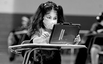A young girl wearing a mask sits at a desk with an iPad.