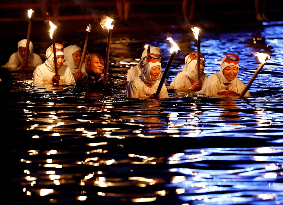 A line of people wearing white clothing and swim masks wades in deep water at night, carrying torches.