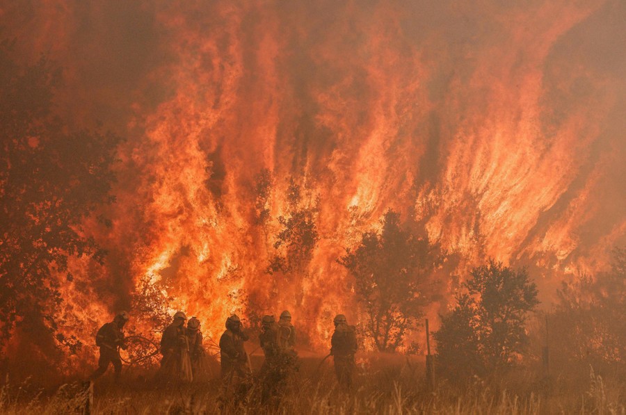 About eight firefighters stand together in a field in front of a huge wildfire.