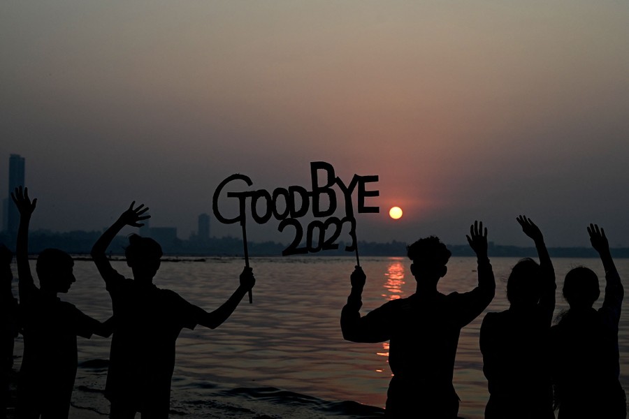 People stand on a beach, waving at the setting sun and holding a sign that reads "Goodbye 2023."