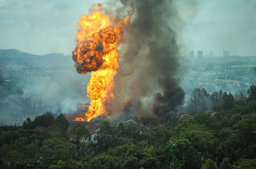 A column of fire erupts into the sky from a crater in a wooded residential area.