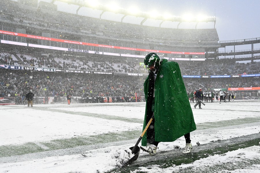 A football player shovels snow on the sidelines of a field in a large stadium.