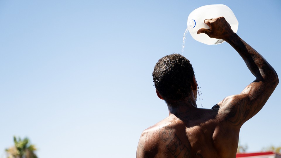A shirtless man seen from the back standing in the sun, pouring a jug of water over his head
