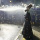 A protester wearing a traditional outfit and gas mask stands in front of a line of riot police, being sprayed in the face with pepper spray.