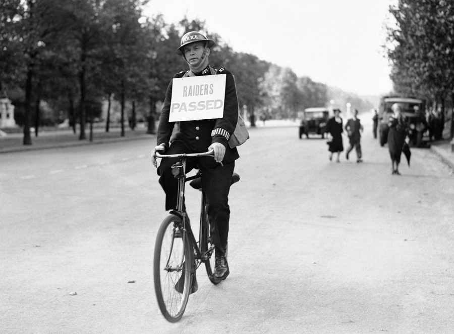 A policeman rides a bicycle down a street, wearing a sign on his chest that reads "Raiders Passed."
