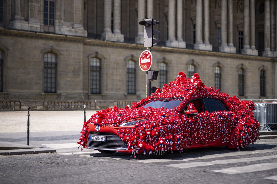 A car that is completely covered with plush versions of the Paris 2024 Olympic mascot, a personified traditional red hat