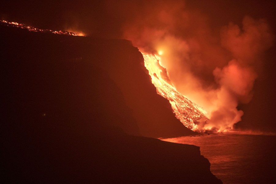 Lava flows over a steep ledge into the ocean, seen at night.