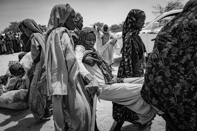 Picture of female relatives carrying Maryam Muktar Khalil, 80, to a truck to be transported by the Tiné Emergency Response Room, part of Sudan’s mutual-aid movement, to a transit camp in eastern Chad