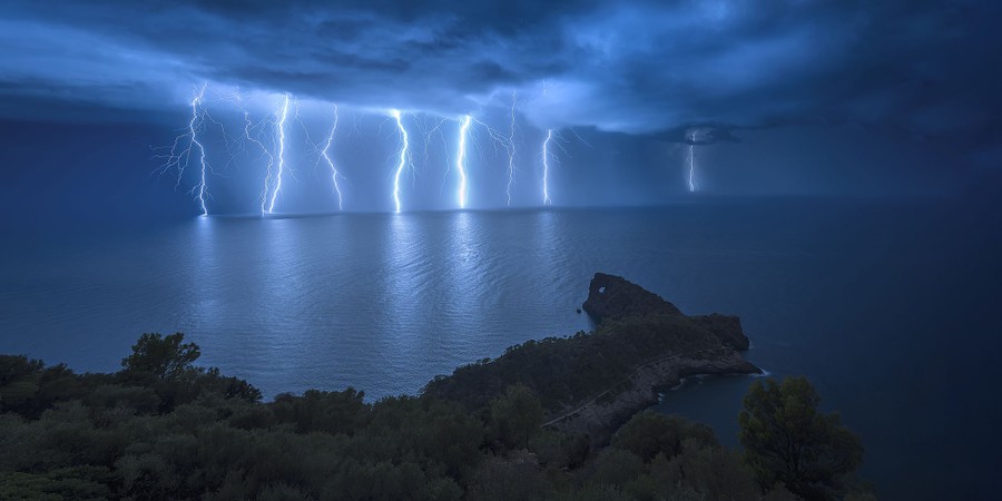 Multiple lightning strikes appear over the ocean, far from a rocky point at night.