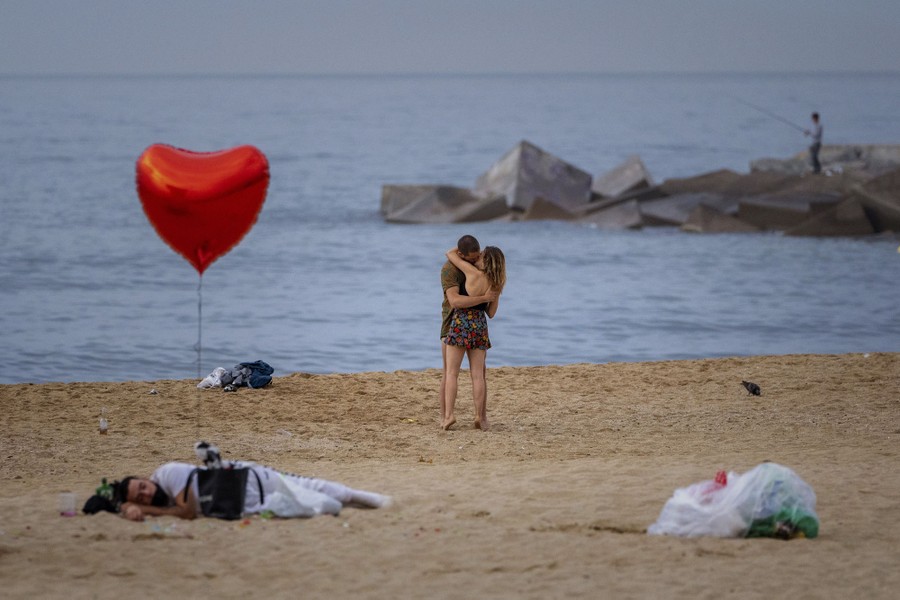 A couple kisses on a beach while, nearby, a person lies on the sand with a heart-shaped balloon tied to their bag.
