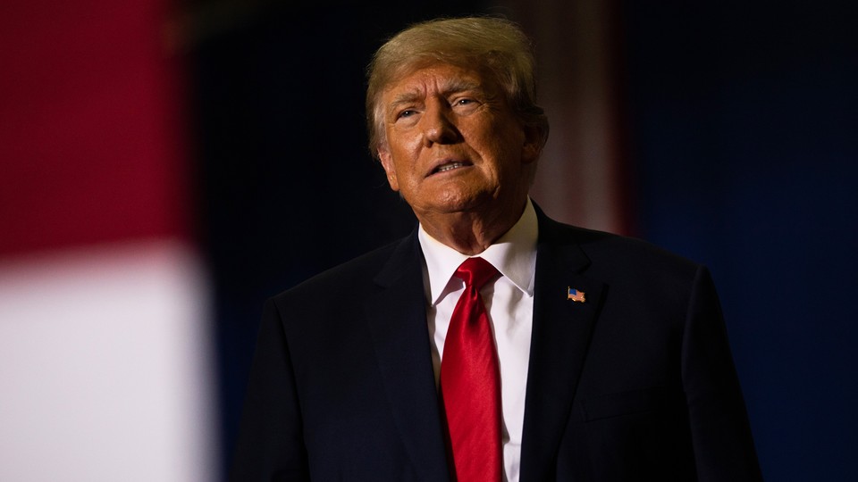 Color photograph of Donald Trump talking, wearing a black suit with a red tie.