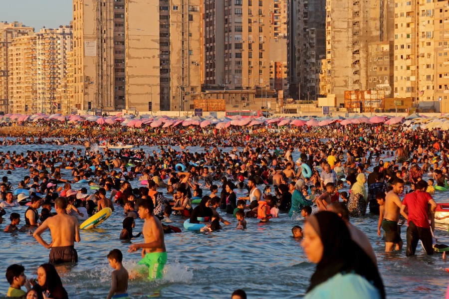 Hundreds of people gather and play along a beach near tall buildings.
