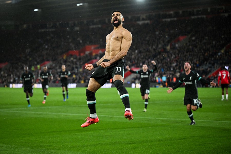 A shirtless soccer player jumps in celebration as his teammates run toward him on a soccer pitch.