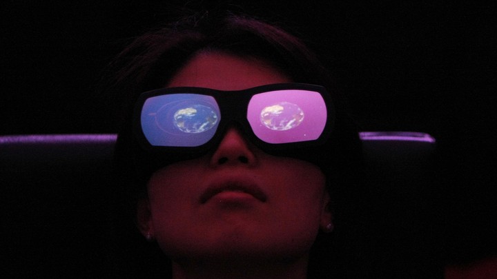 A person wears large glasses with one red and one blue lens and an image of the Earth reflected in each.