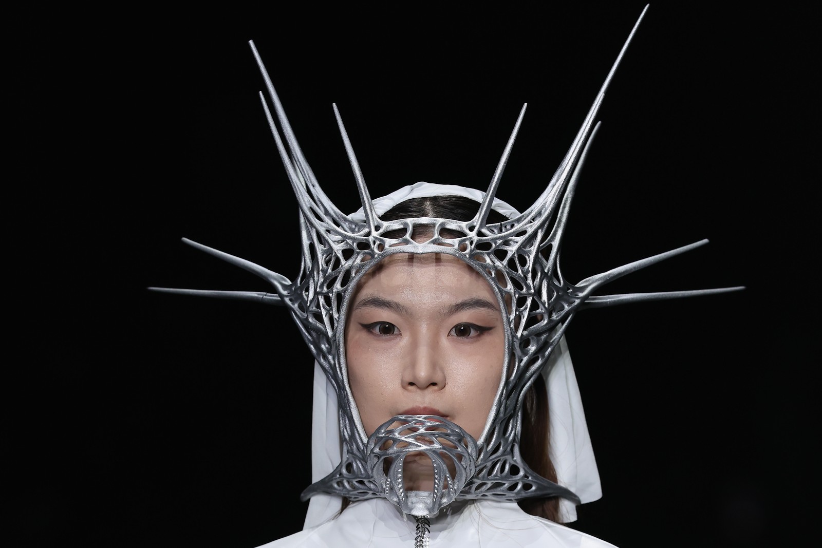 A model walks a runway, wearing a spiky metal headdress that circles their face.