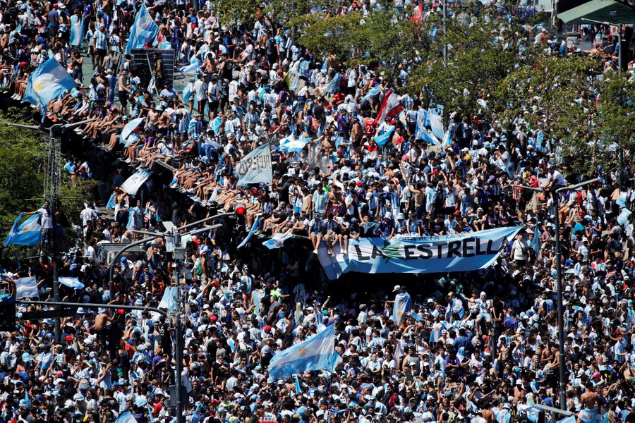 Hundreds of soccer fans sit and stand along a city street.