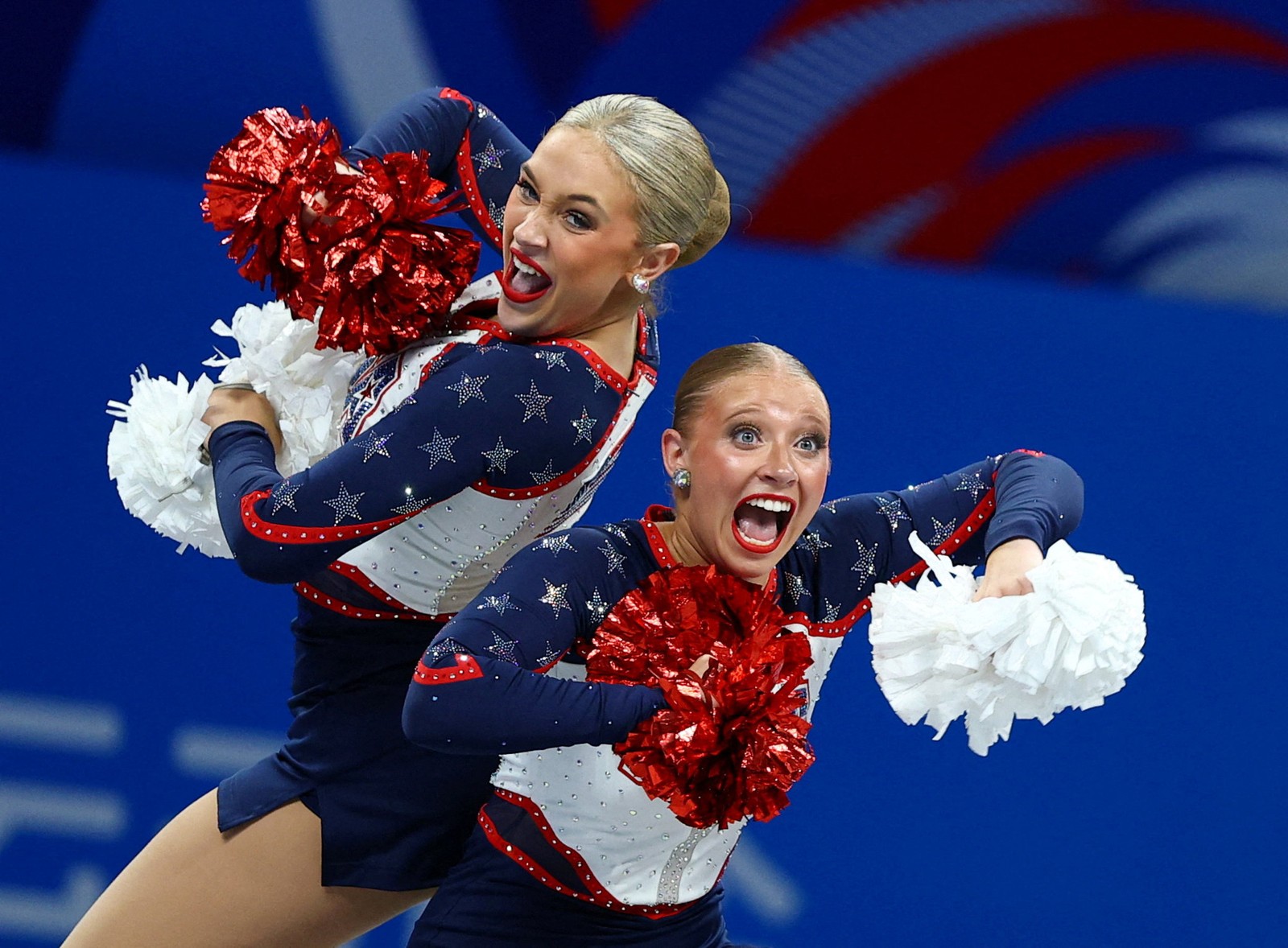 Two gymnasts holding pom-poms, seen smiling broadly during a competition.