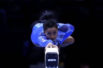 The gymnast Simone Biles lies stretched-out atop a balance beam wearing a shiny blue leotard