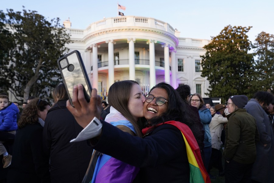Two people take a selfie as they kiss in front of the White House, in a crowd.