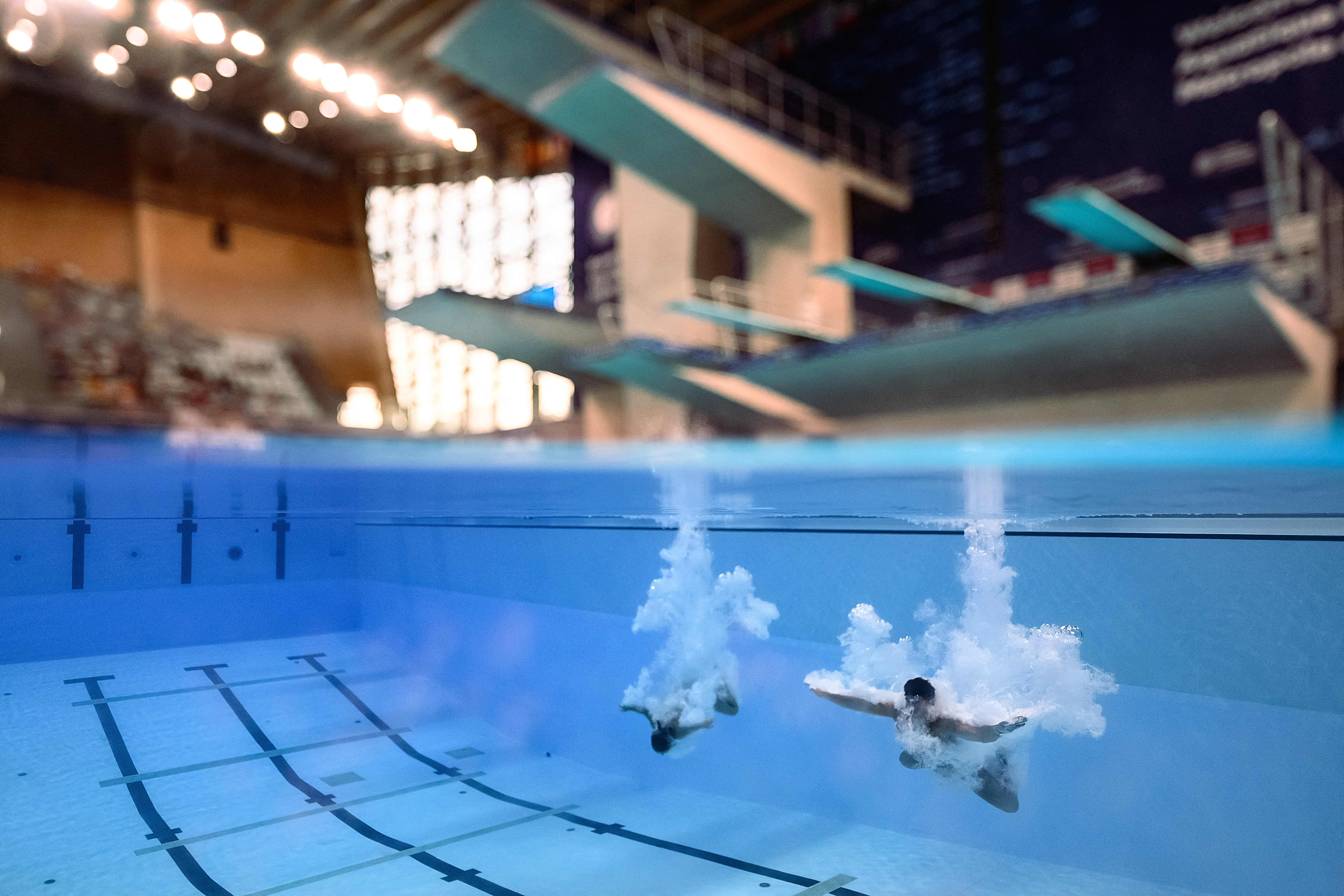 Two divers, seen just below the surface of a pool, surrounded by bubbles after a dive