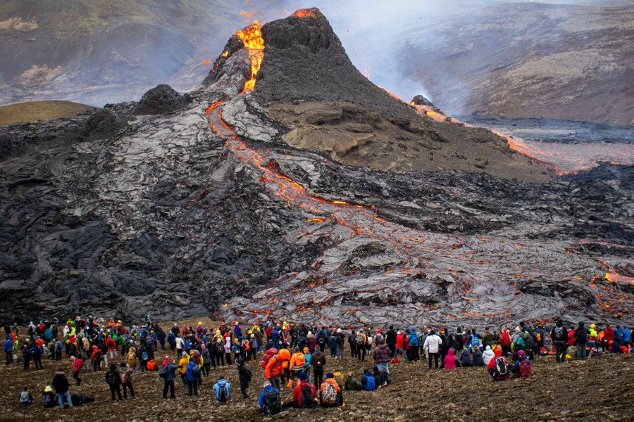 Dozens of people sit on a hillside looking toward a small volcano with lava erupting out of the crater at its peak.