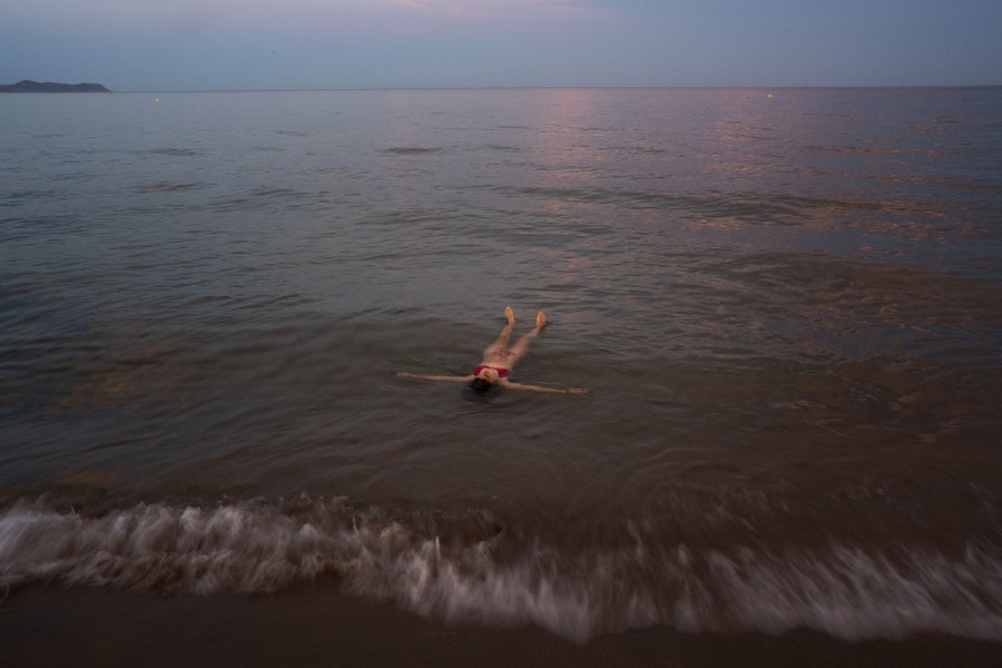 A swimmer floats on their back in shallow water near the shoreline.