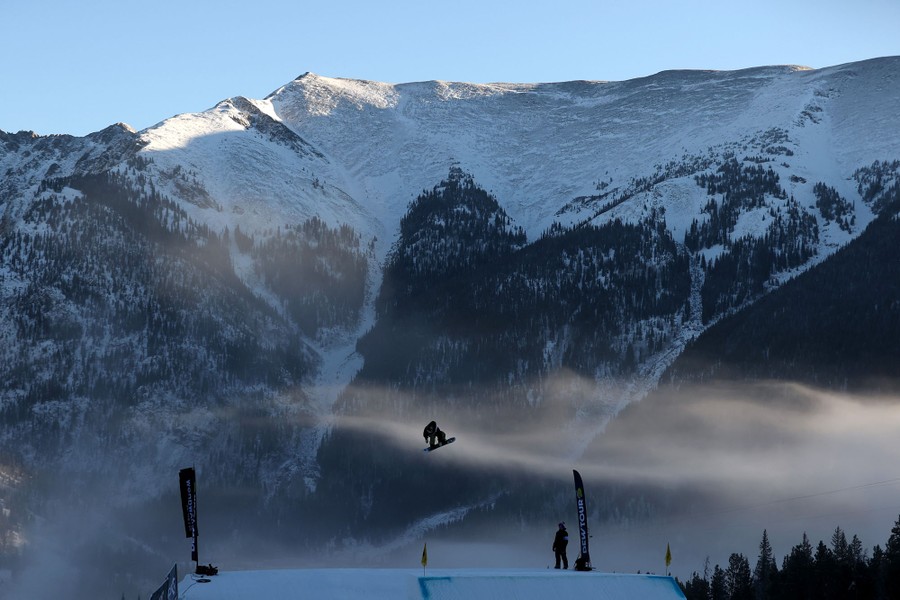 A snowboarder makes a jump, seen in front of snow-covered mountains.