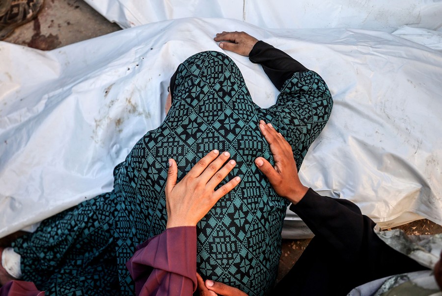 Mourning people place their hands on the back of another, who lays their head on a body that has been covered with a plastic sheet.