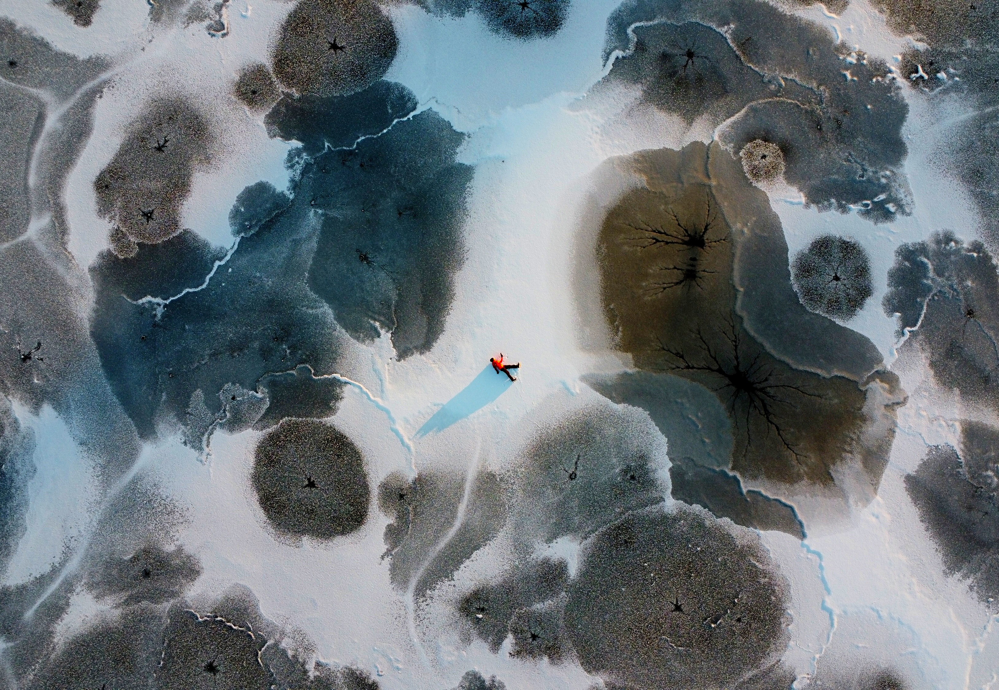 A top-down aerial view of a person wearing cold-weather gear who is lying on their side in a 'walking' pose on a frozen lake