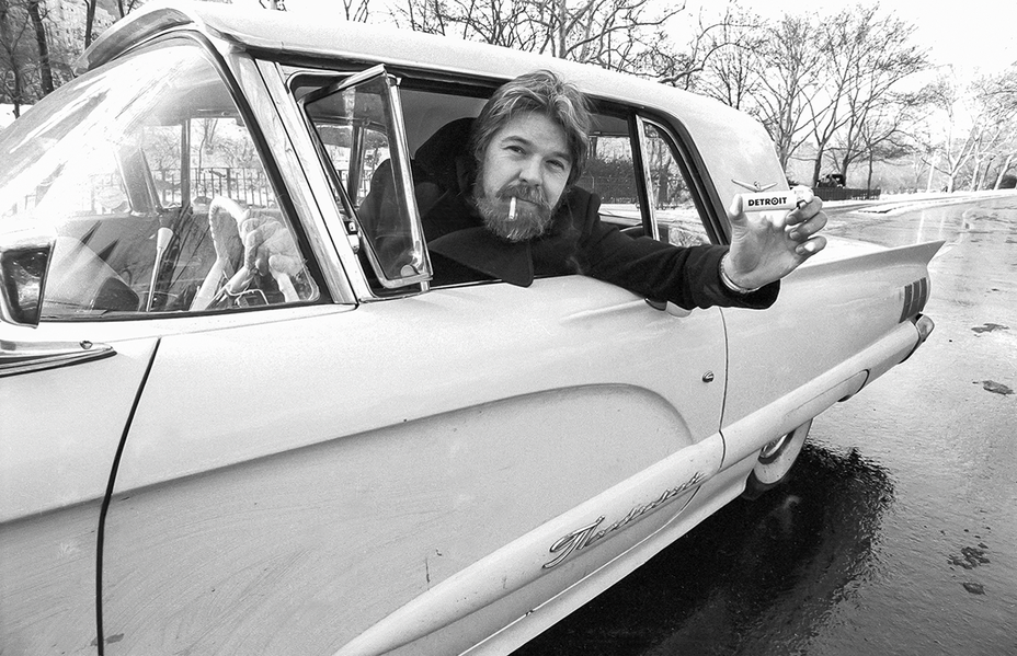 black-and-white photo of man with beard and cigarette in mouth holding item labeled DETROIT out window while driving vintage car