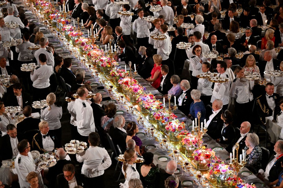 Many members of a wait staff carry trays of desserts along a very long table at a formal banquet.