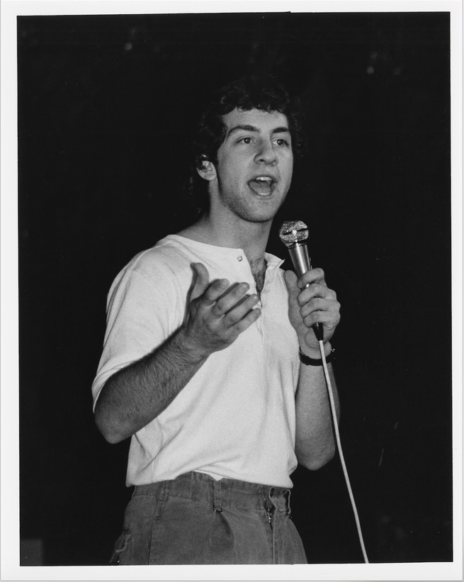 black-and-white photo of dark-haired man in white henley and jeans speaking into wired microphone and gesturing