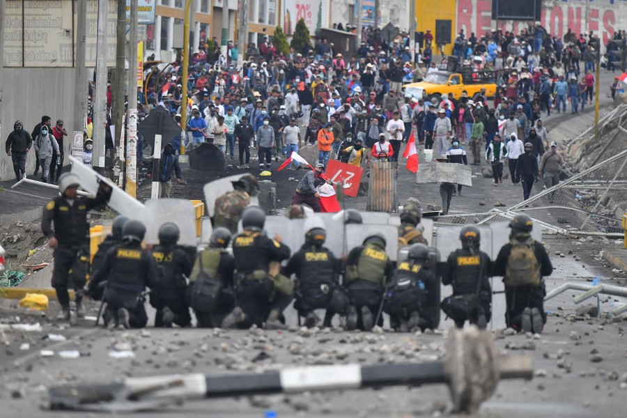 A group of protesters stands in a street, facing off against a line of riot police holding shields.
