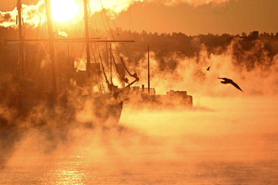 A sunrise lights up mist above water and a moored ship.